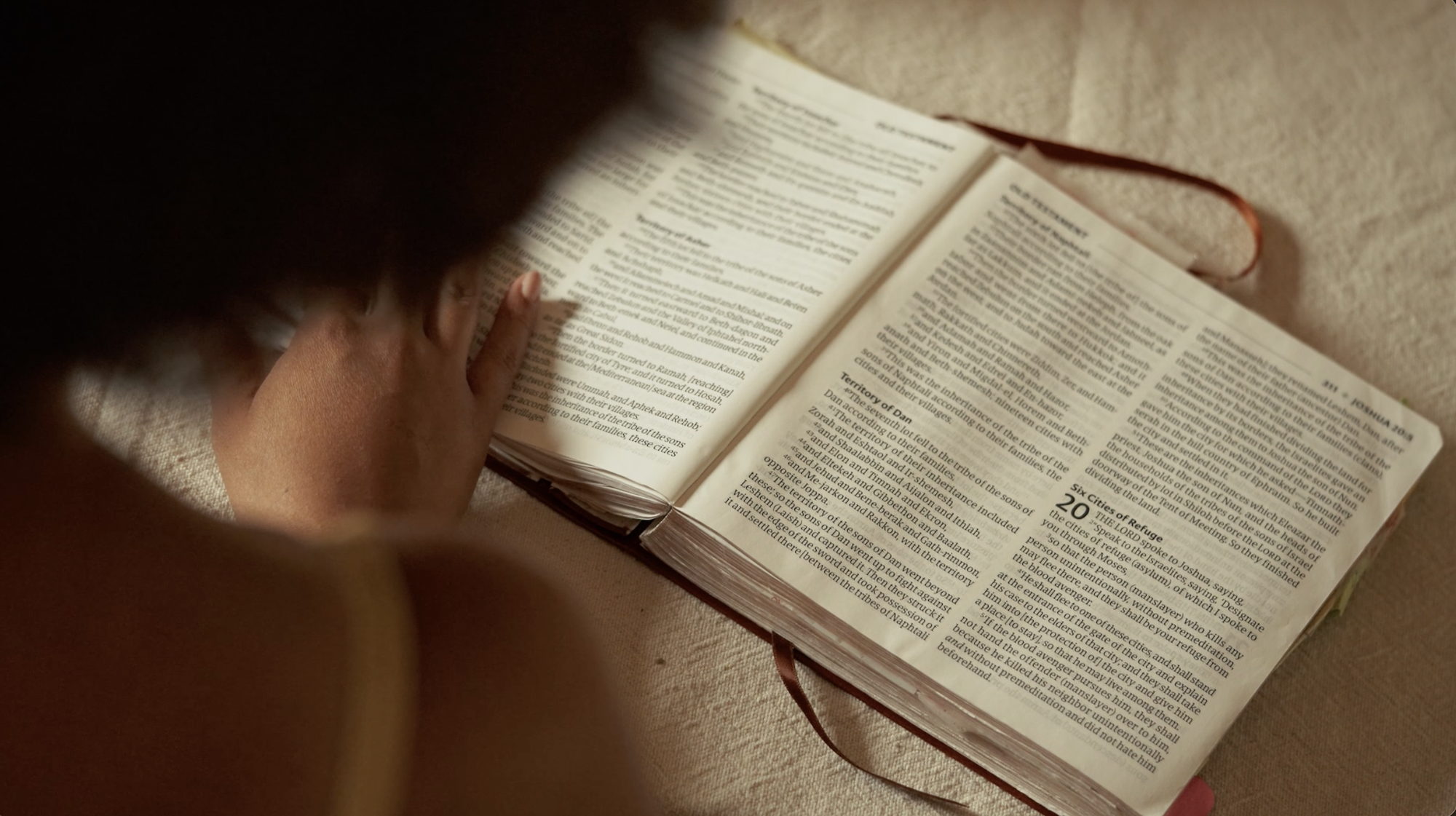 Over her shoulder, an open Bible on the table in window light.