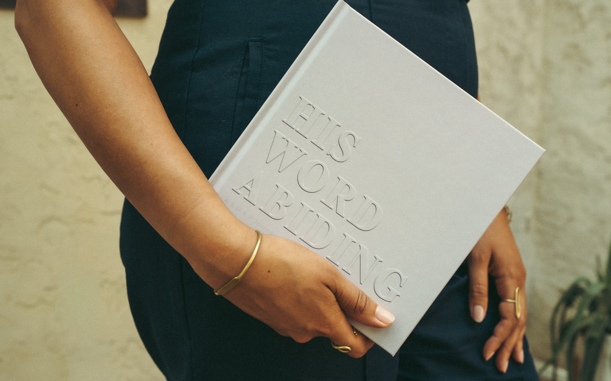 A woman in a navy jumpsuit holds the His Word Abiding journal at her side, its embossed cover catching warm light.