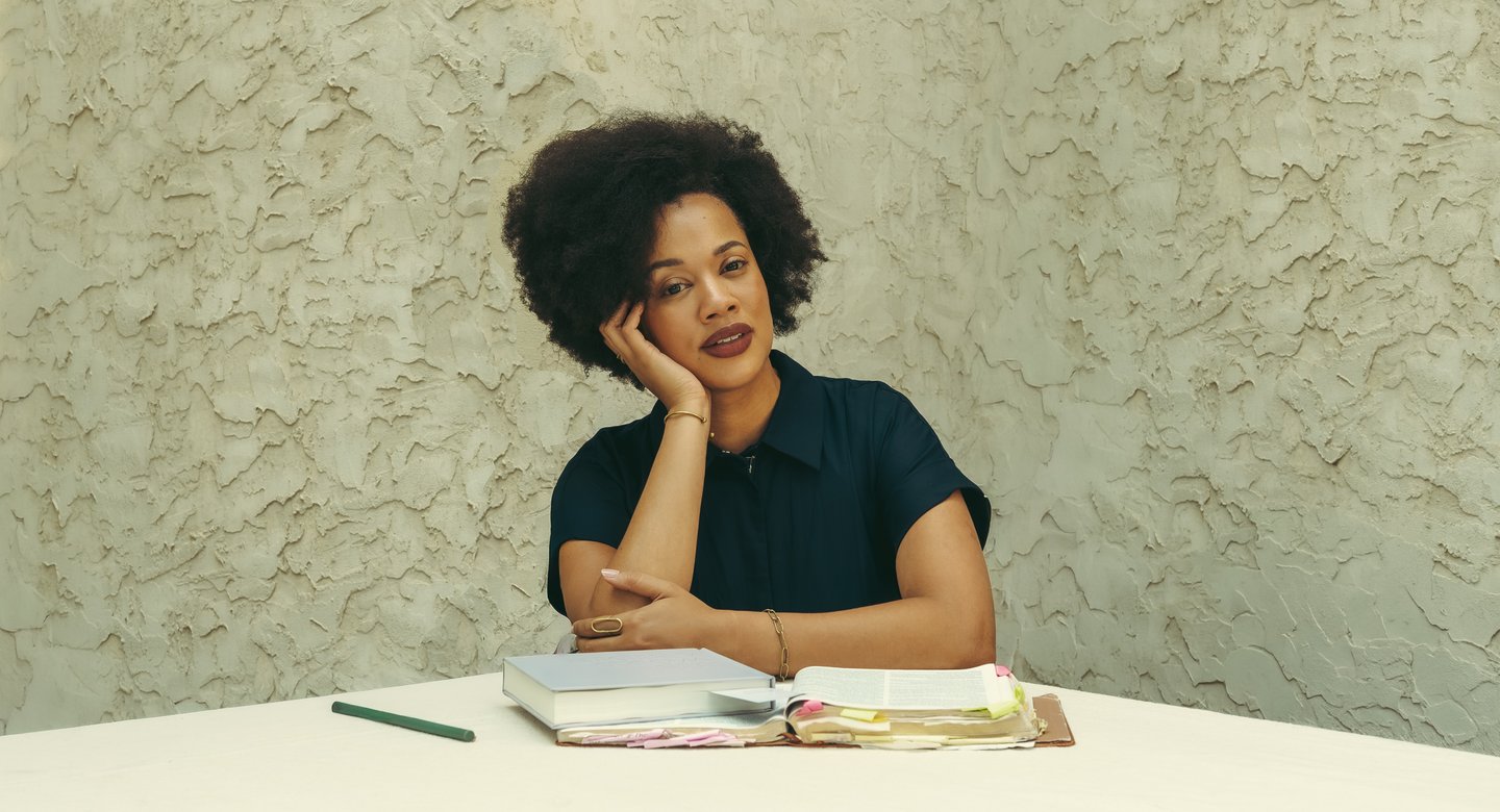 Lauren, founder of Wellness in Christ, seated at a table with her chin resting in her hand and an open journal before her, against a warm plaster wall.
