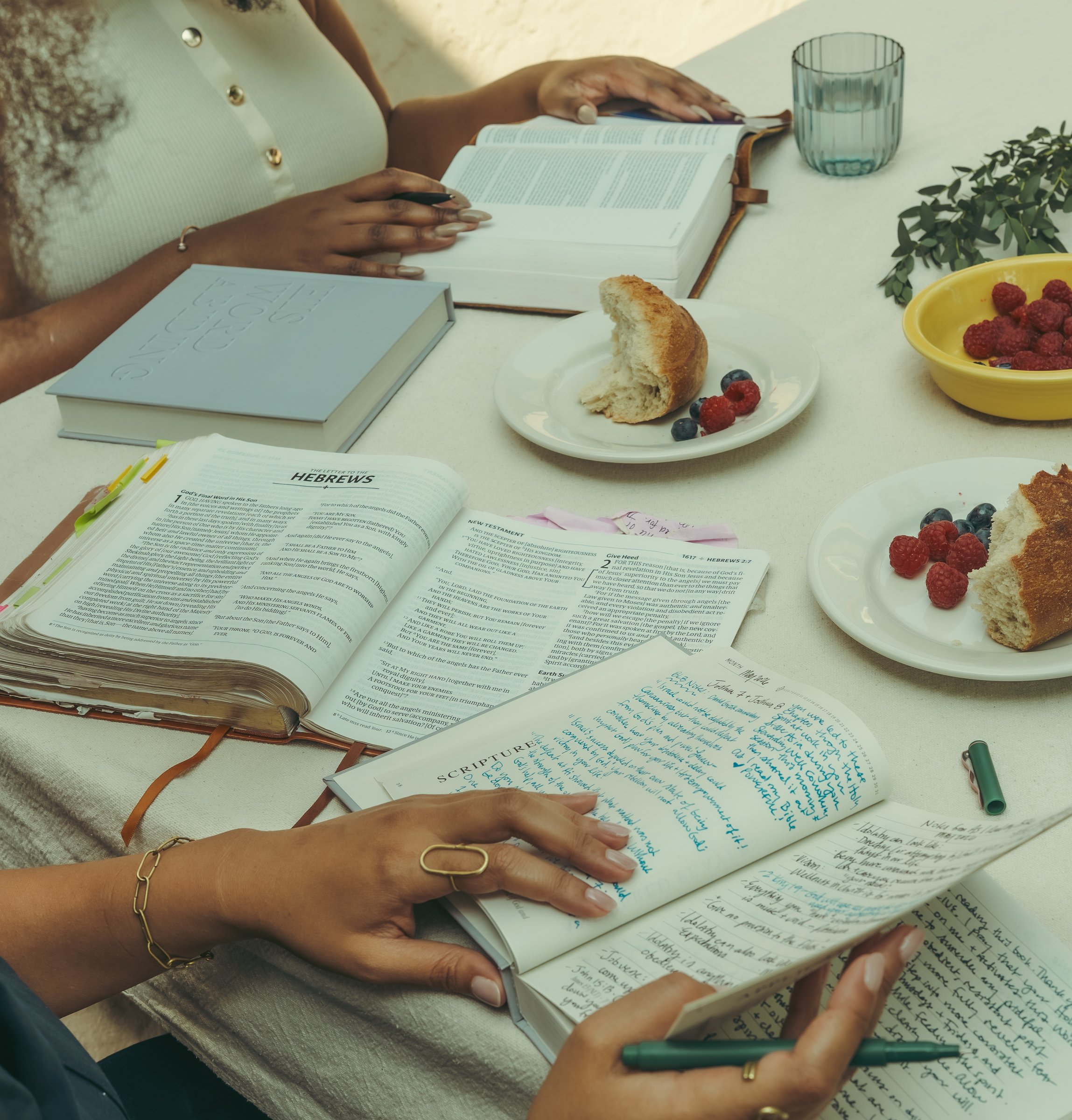 Seen from above, women's hands rest on open Bibles and a handwritten journal at a table set with bread and berries.