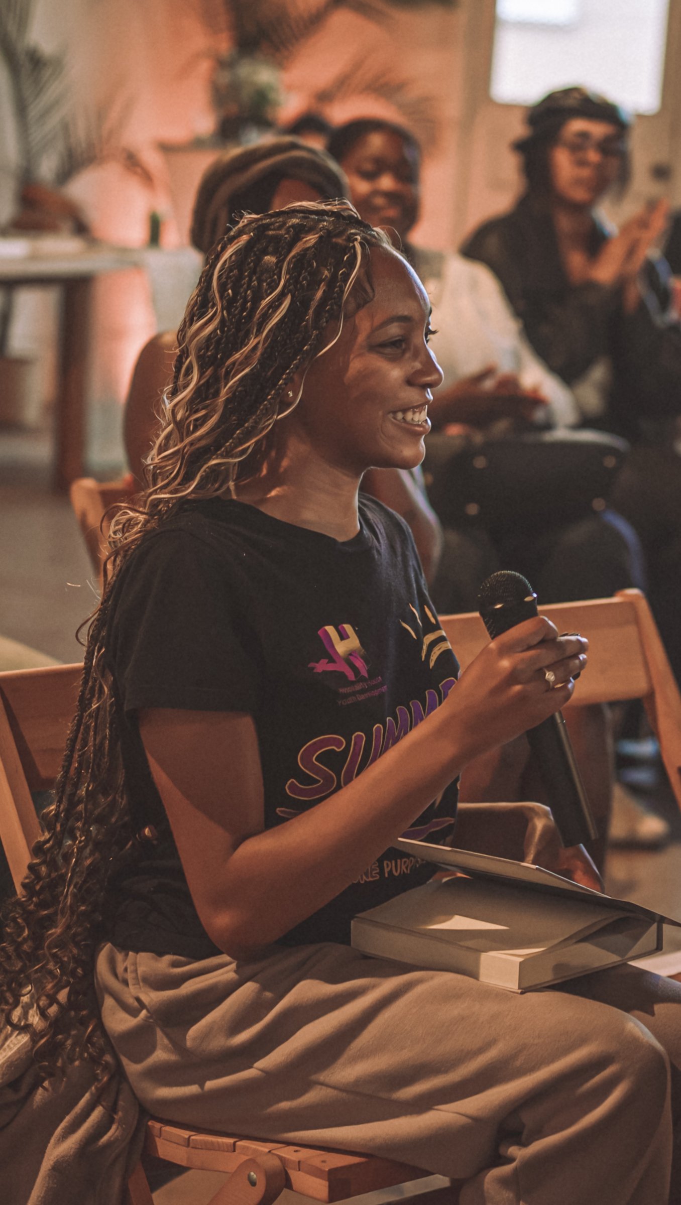 A young woman with long braids smiles mid-sentence, microphone in hand and a journal resting on her lap.