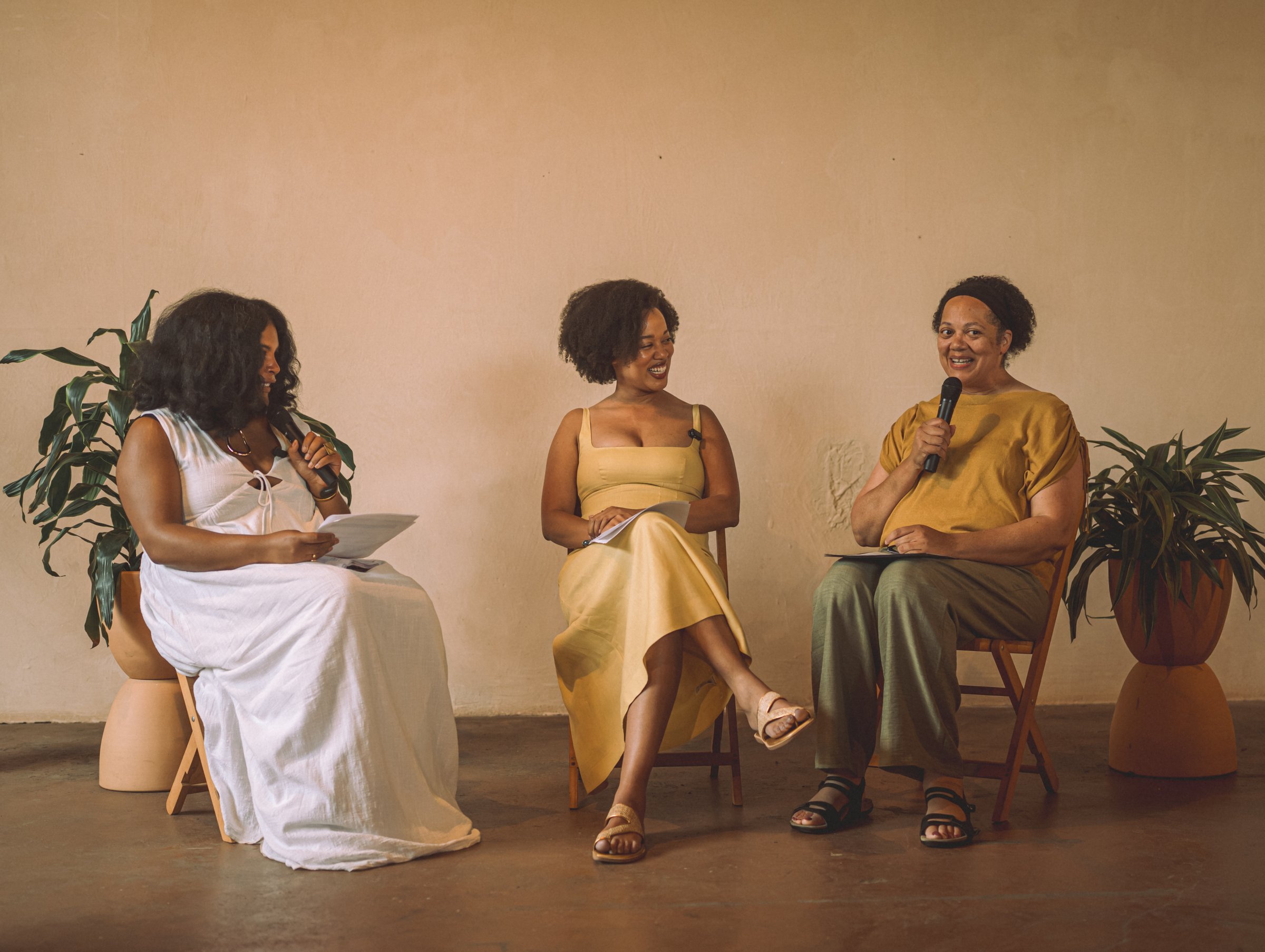 Three women in conversation on chairs before a warm plaster wall: one in a white dress, Lauren in a yellow dress, and one in a mustard top holding a microphone.