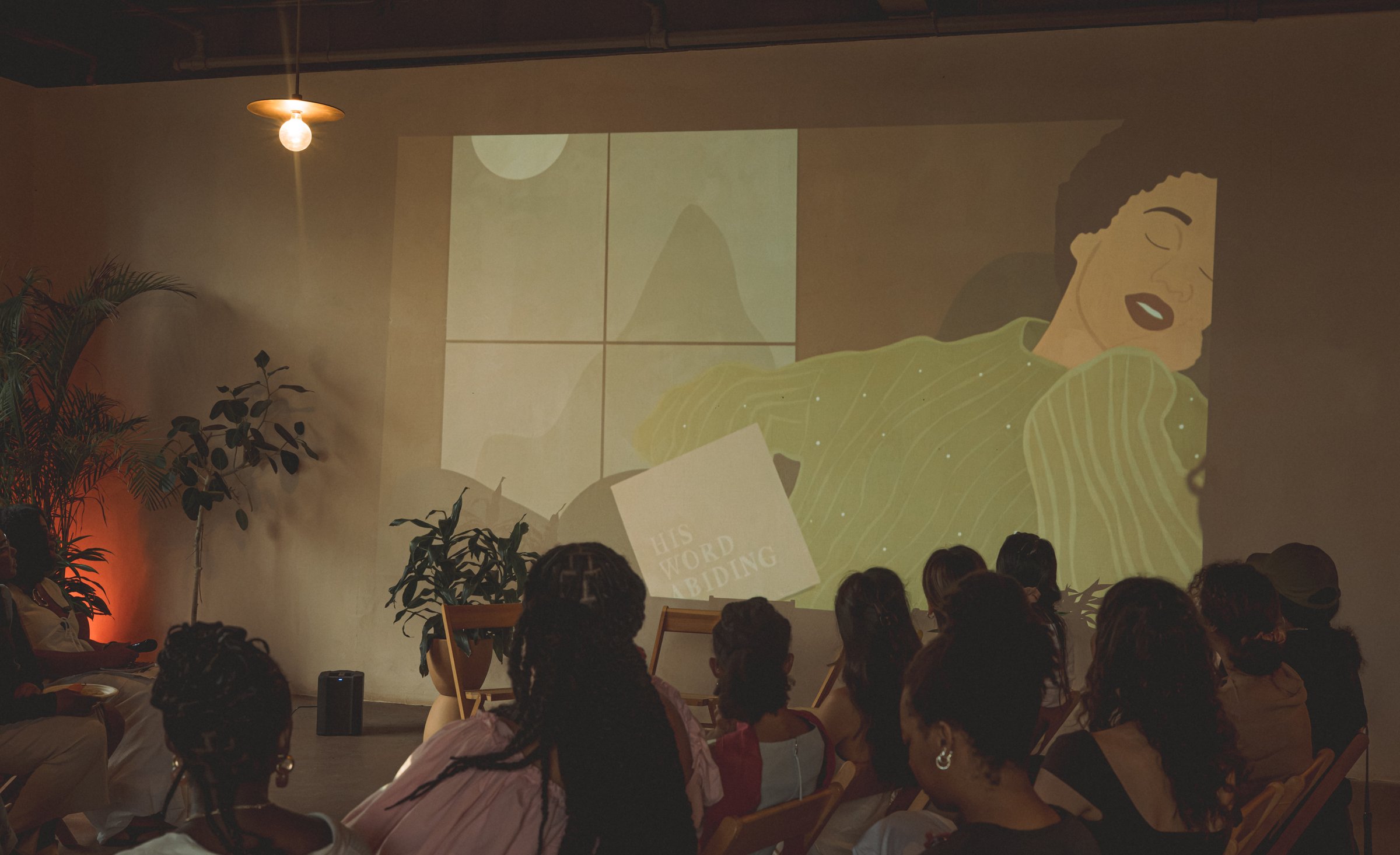 Guests seated in a dim room face an illustration projected on the wall, a single lamp glowing overhead.