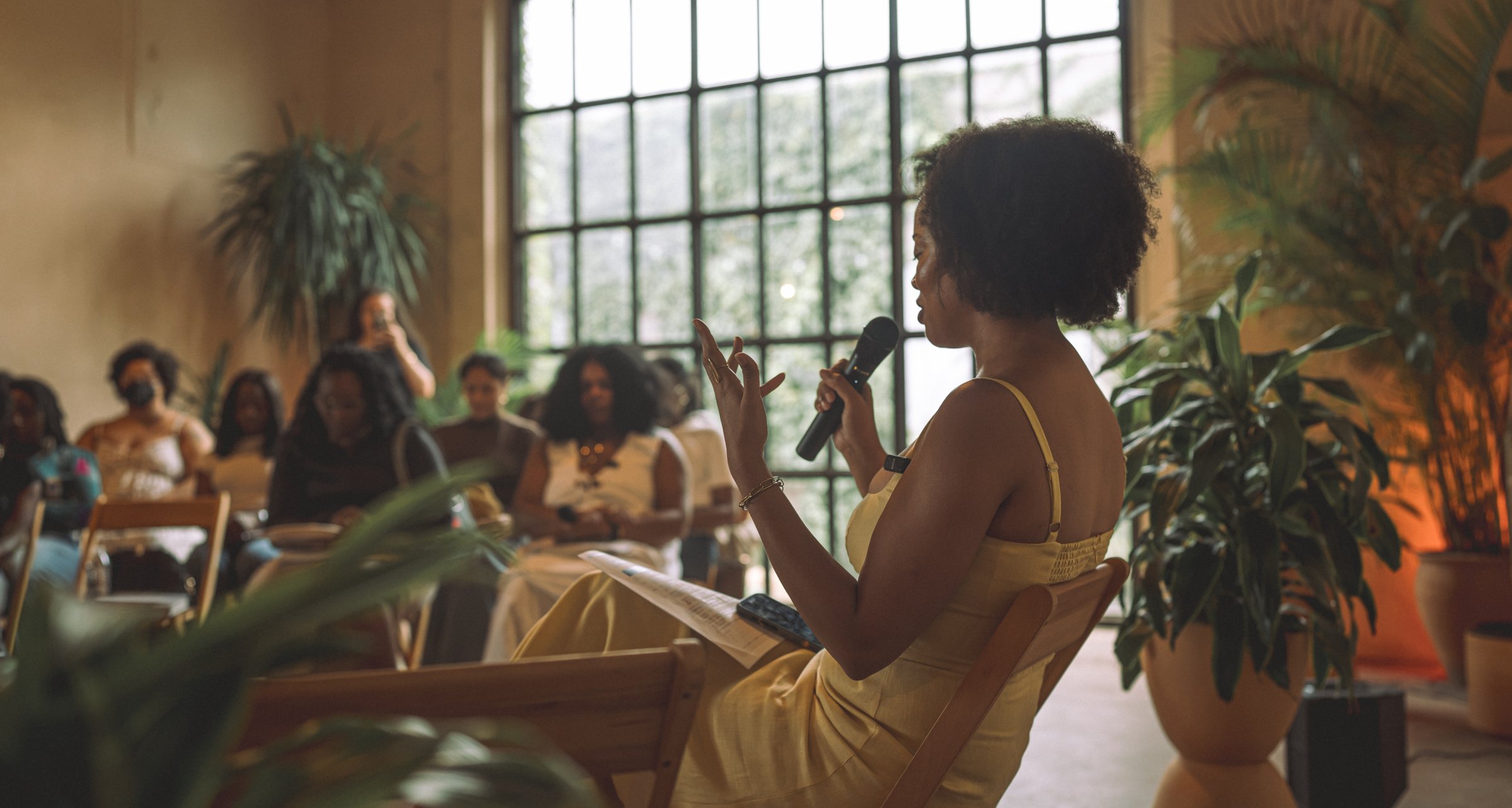 Lauren, in a yellow dress, sits before the room with a microphone and her eyes closed in prayer, guests and tall windows behind her.