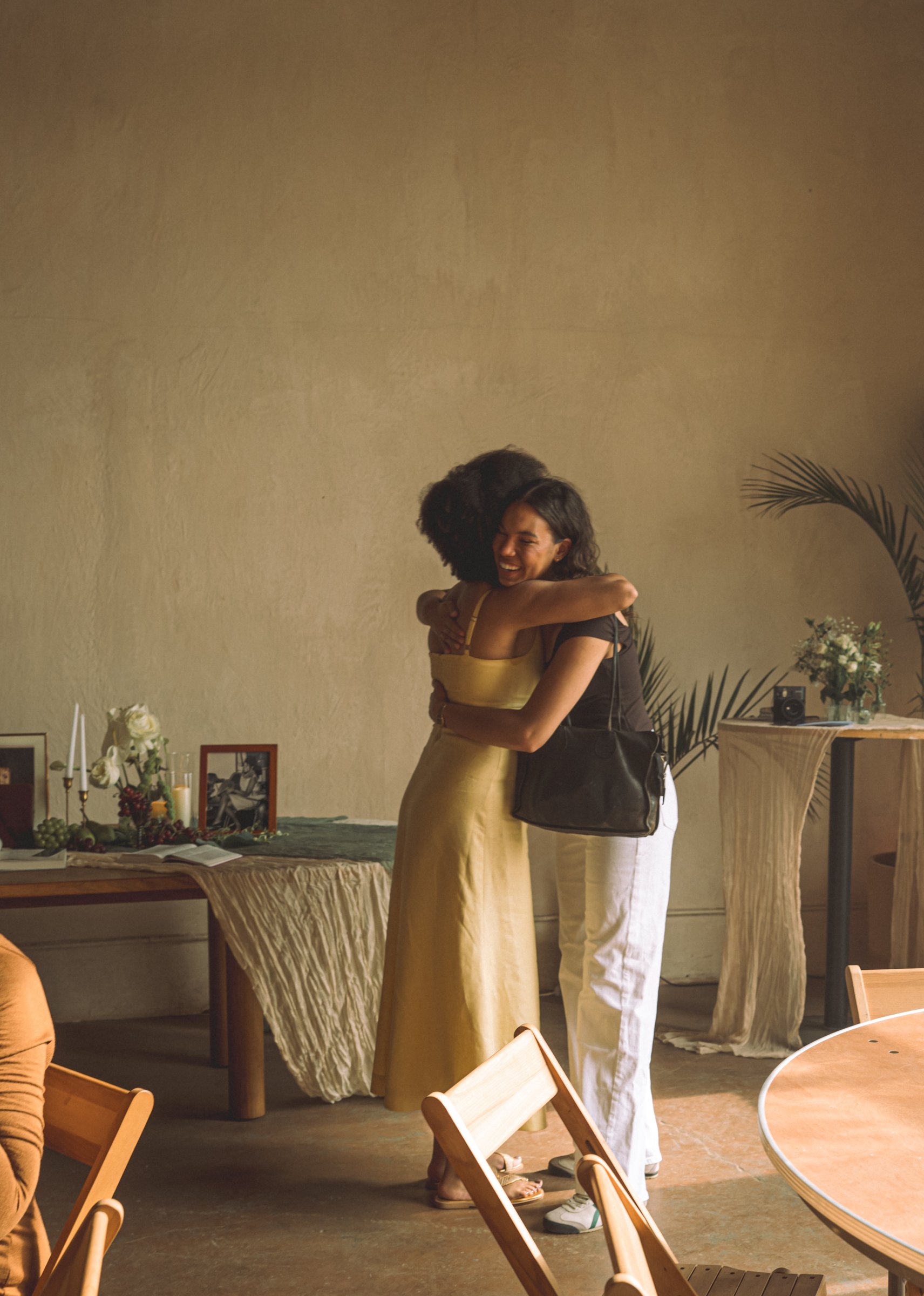 Lauren, in a yellow dress, embraces a friend in white beside tables set with candles and flowers.
