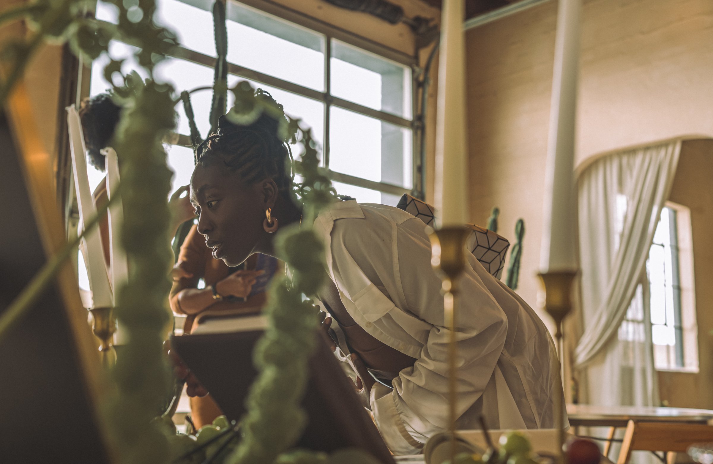 Seen through trailing plants, a woman sits by a tall window in quiet thought while a guest in a white head wrap leans in nearby.