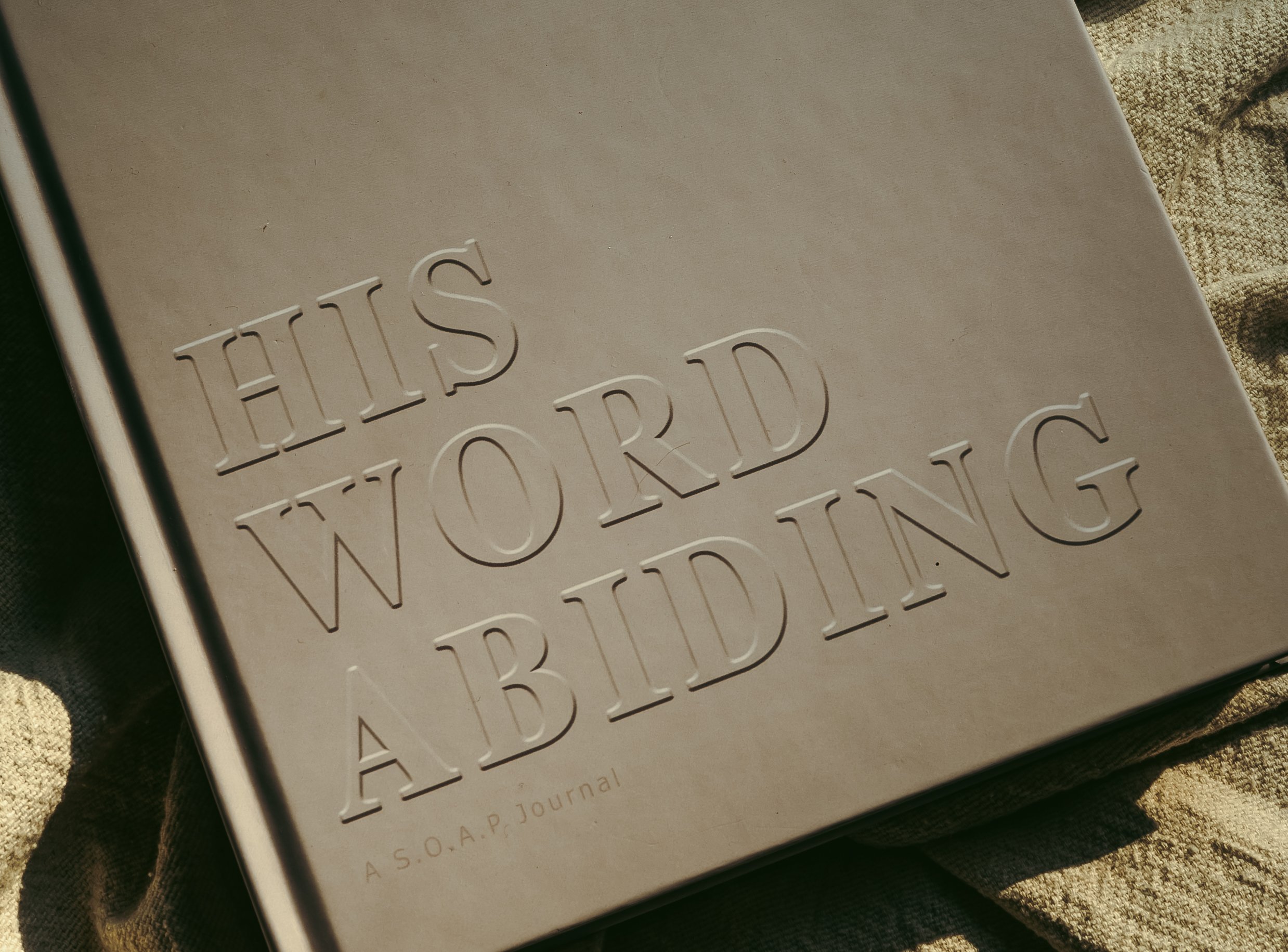 The His Word Abiding journal, its navy cover embossed with the title, resting on soft linen in warm light.